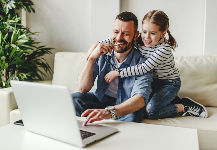A child with her arms around her father while he is working at a computer