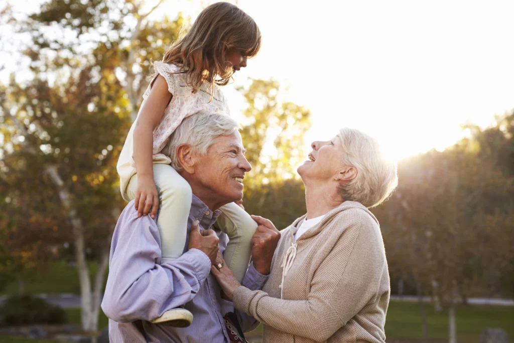 Grandparents smiling and laughing outdoors with young granddaughter sitting on grandfather's shoulders
