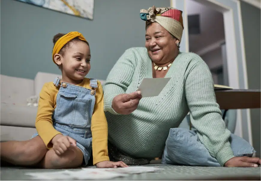 A grandparent sitting on the floor with a grandchild while smiling and looking at papers