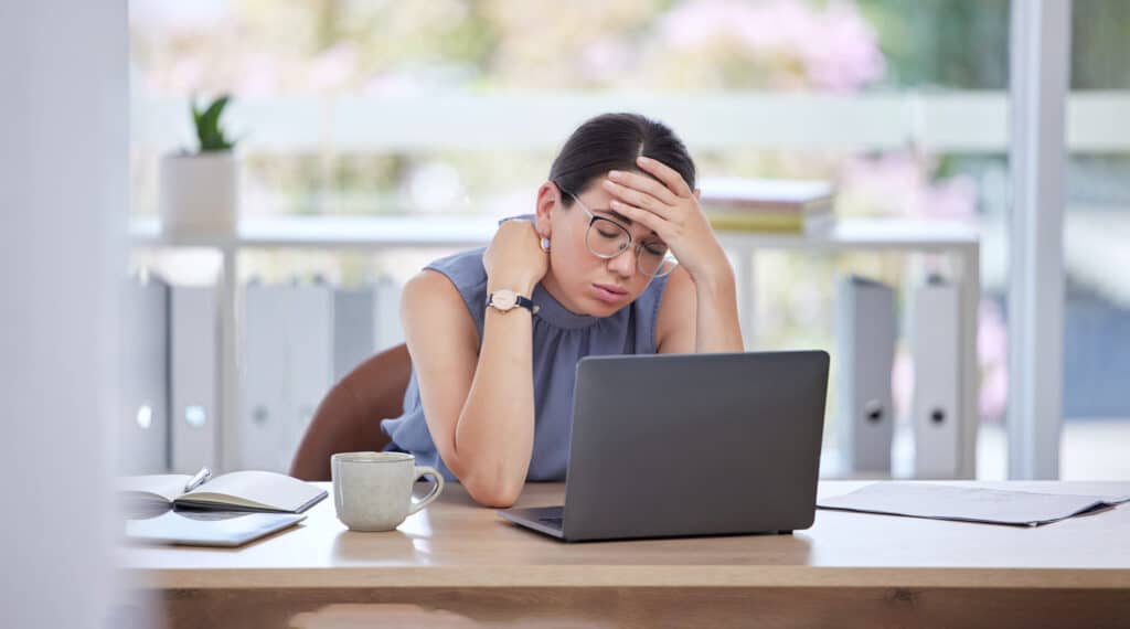 Stressed woman holding her head while looking at a laptop at her office desk with coffee and notebook nearby