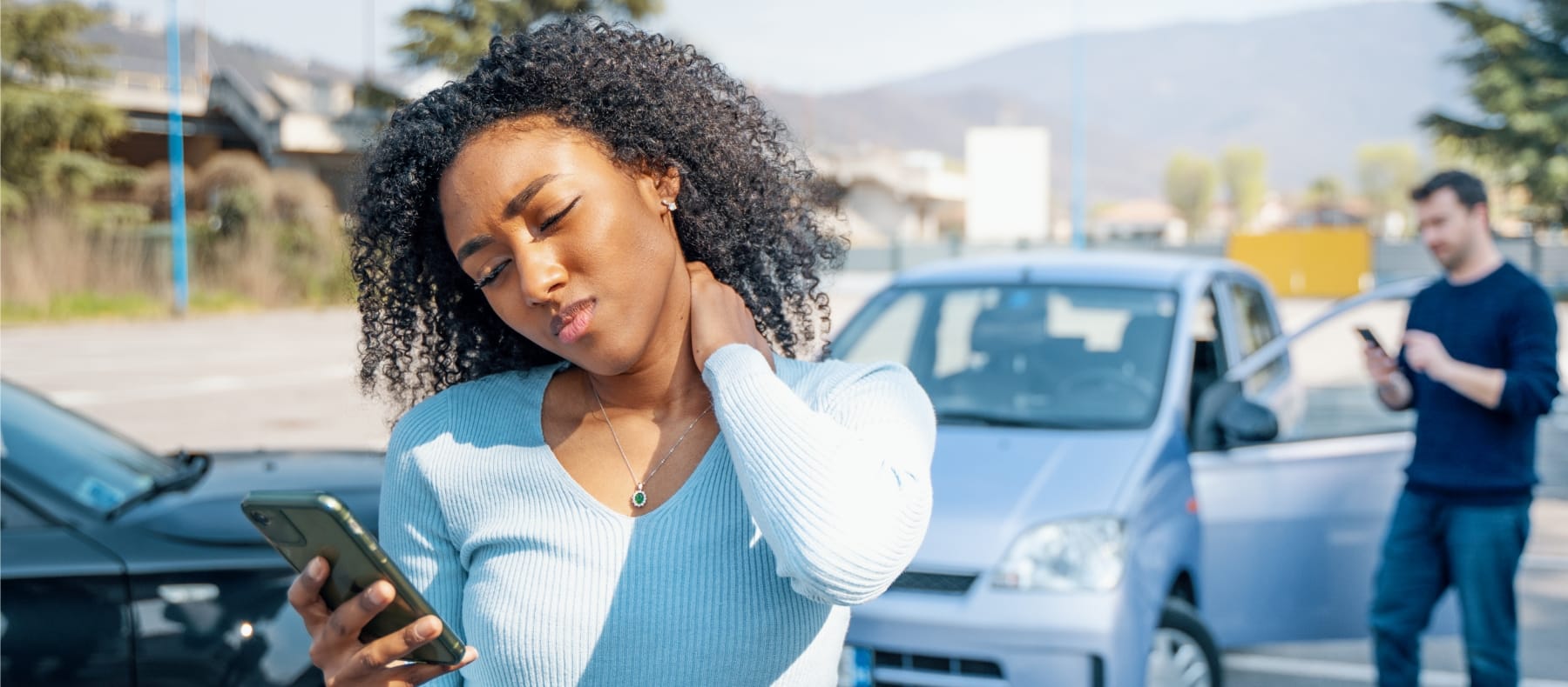 A woman standing near the scene of a car accident, holding her neck while looking at her phone.