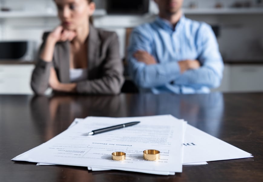 A couple at odds sitting with folded arms. In the foreground lies paperwork and wedding rings