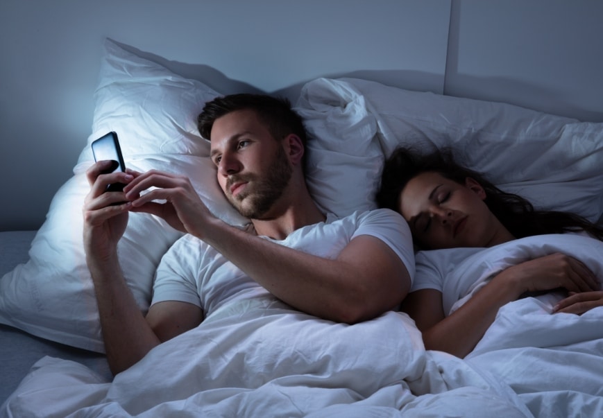 A couple in bed while the wife is sleeping and the husband is on his phone