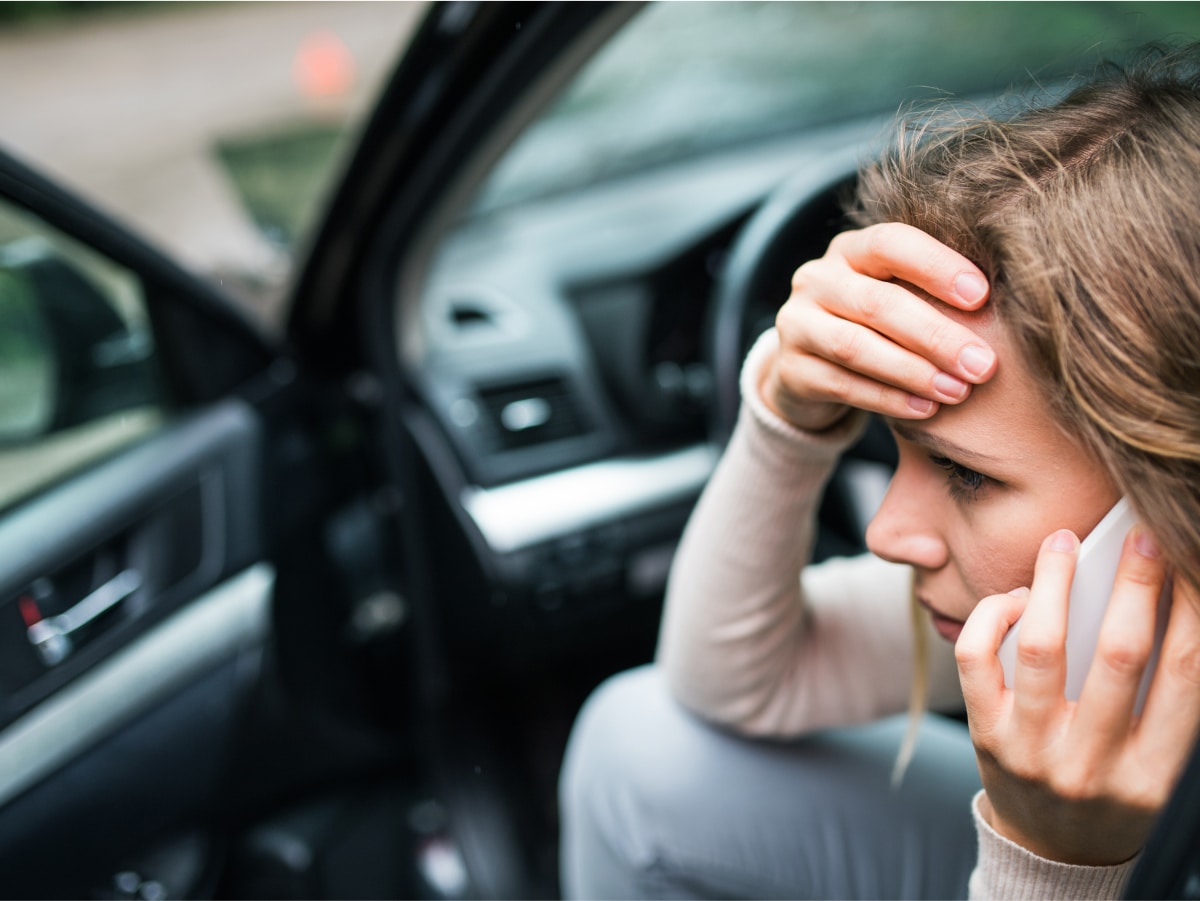 What Should I Do After a Hit-and-Run Crash? 30 A woman with a concerned expression holding her forehead and talking on a cell phone. She is in the driver's seat of a car with the door open.