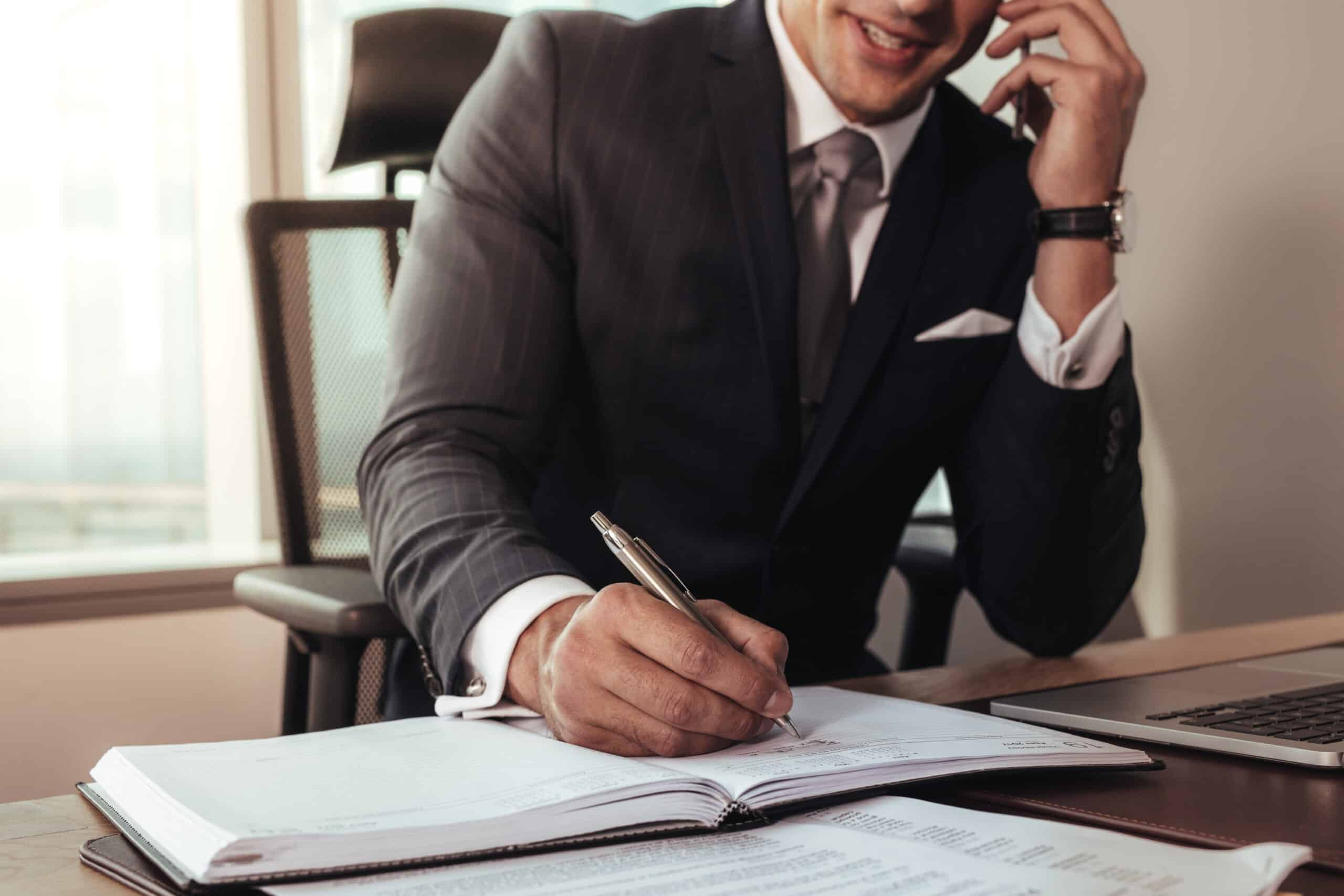 What Should You Expect When You Contact a Lawyer in Charlotte? 15 A businessman in a suit writing notes in a planner while speaking on the phone at his office desk, representing professional communication and business planning.
